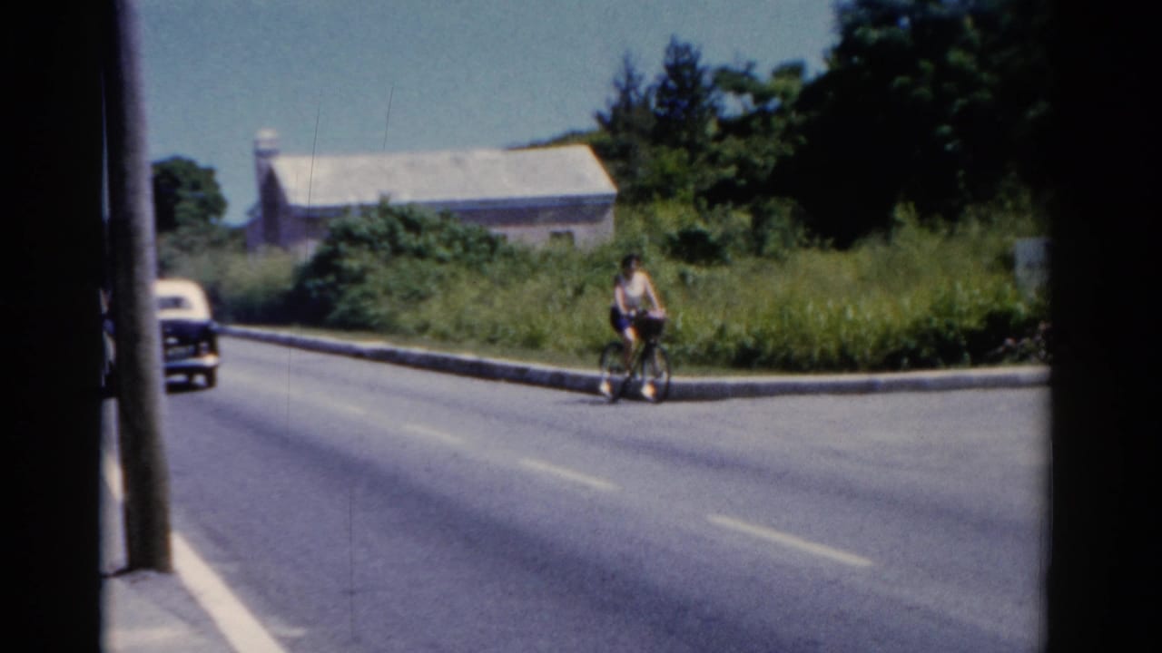 Woman riding a bicycle on a quiet road as a dark-and-light car appears at the left edge.