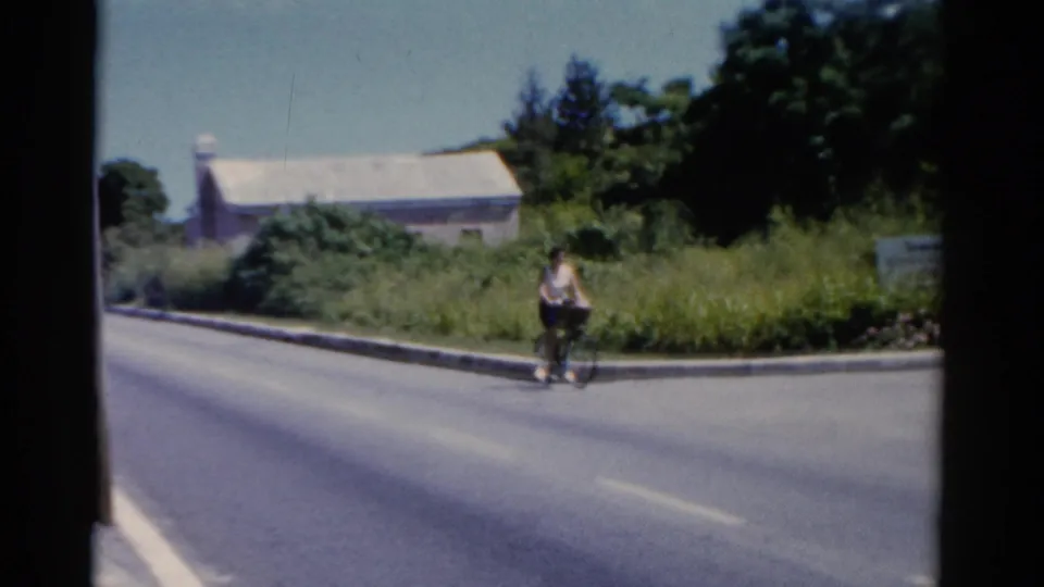 1975 Florida home-movie frame of a woman cycling toward the camera beside a grassy roadside and a light-colored building.