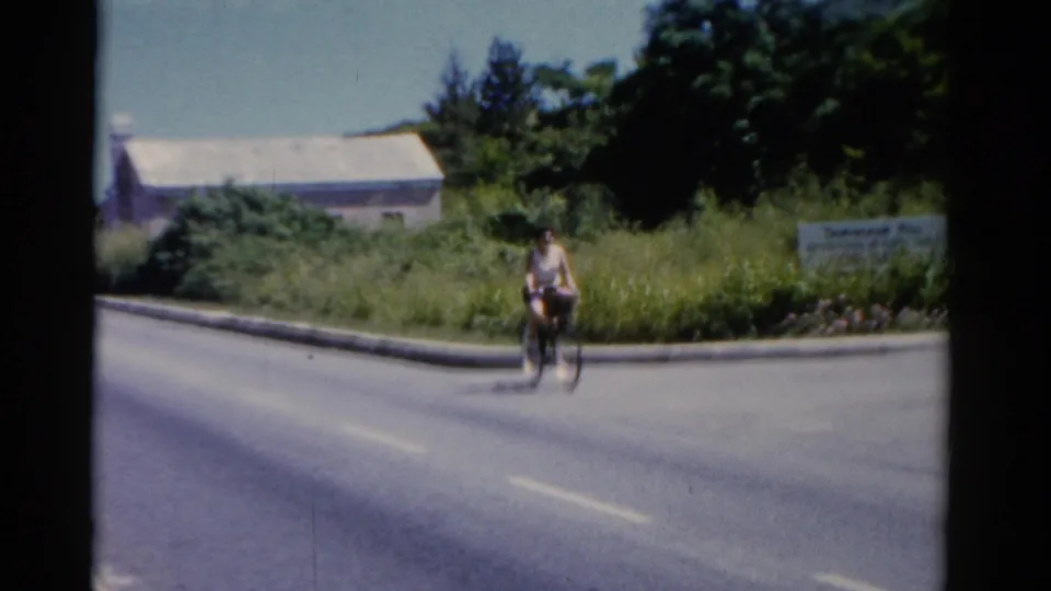 1975 Florida 8mm still with a woman riding a bicycle near the center of the road scene, backed by roadside grass, trees, and a pale building.