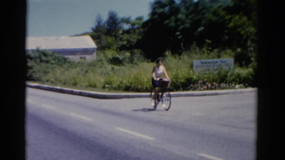 1975 Florida home-movie frame showing the woman cycling past greenery and a pale roadside sign or wall under bright daylight.