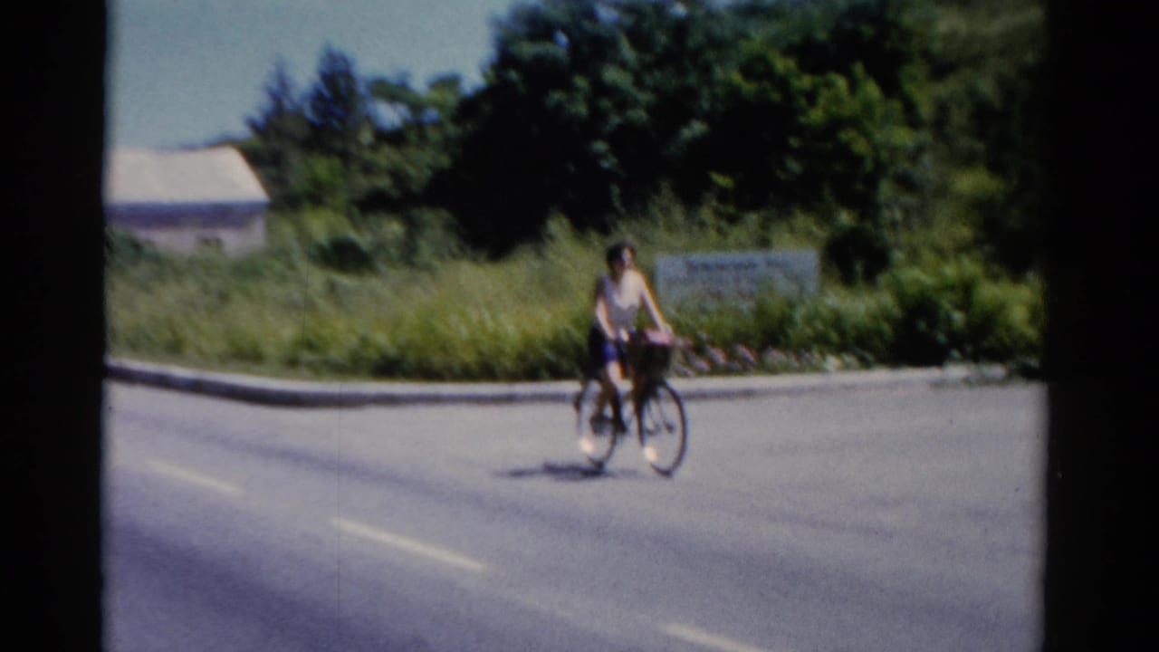 Woman rides a bicycle with a front basket along a Florida coastal road in 1975 home-movie footage.