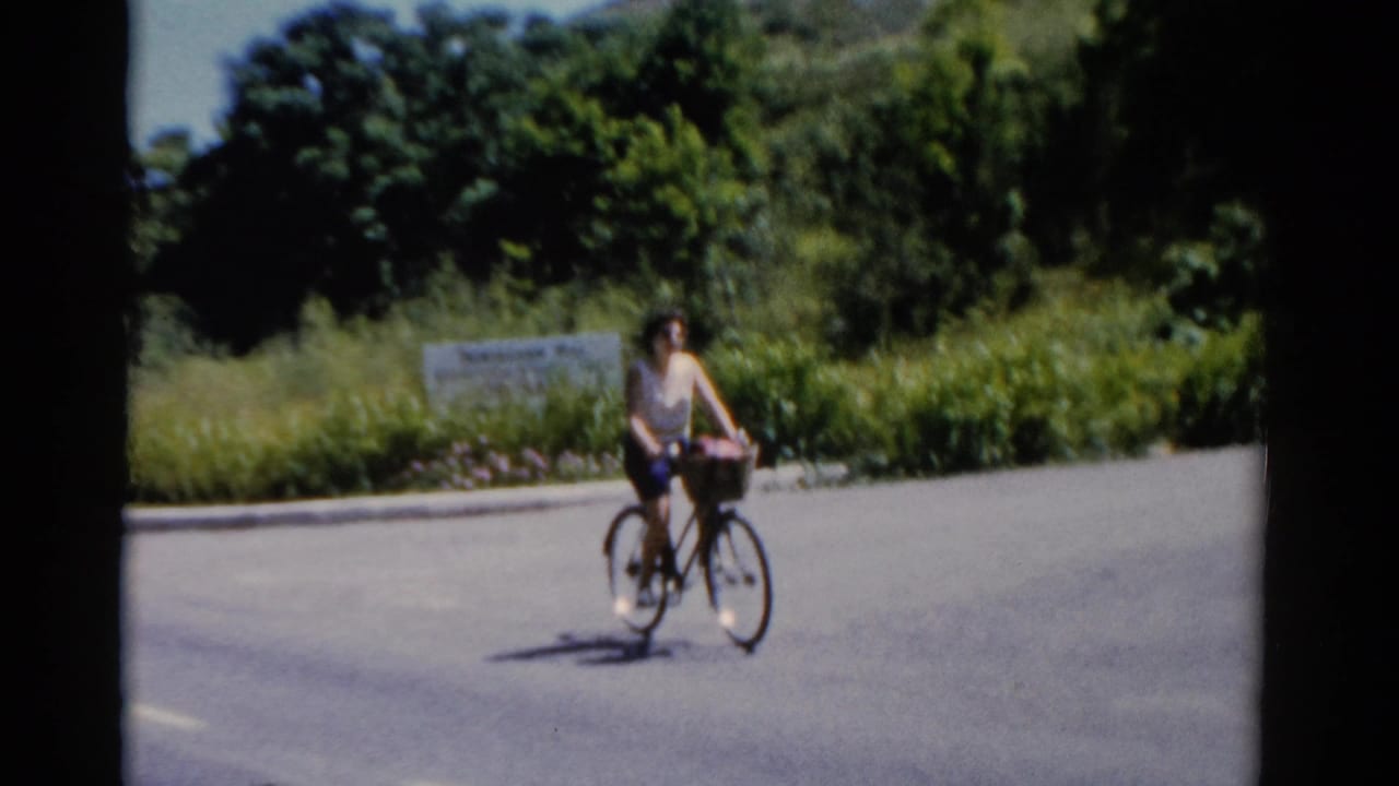 A woman cycles past low roadside planting and a sign in Florida, captured on 8mm film in 1975.