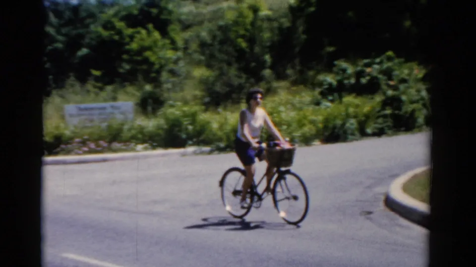 A woman rides a basket-front bicycle near a curb on a sunny Florida road in 1975 home-movie footage.