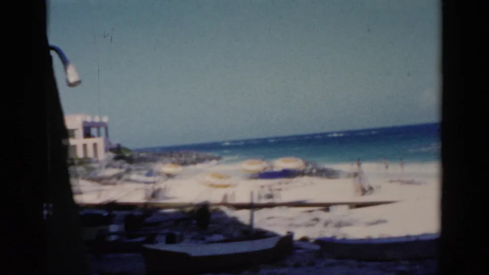 Florida beach view with pale sand, turquoise water, scattered umbrellas, and a small boat-like shape in 1975 8mm footage.