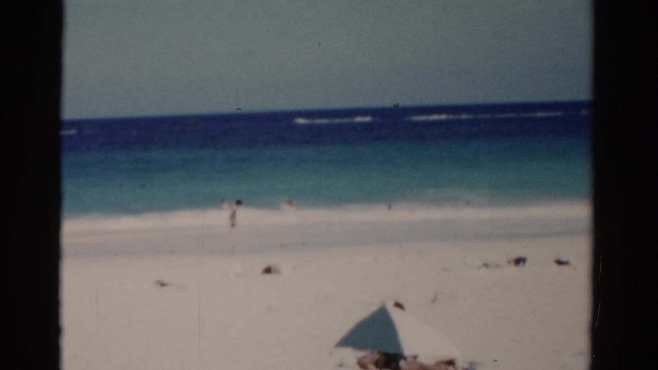 1975 Florida 8mm beach view with white sand, turquoise surf, distant people, and a pale umbrella at the lower edge.