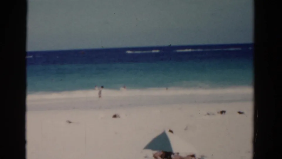 1975 Florida 8mm beach view with white sand, turquoise surf, distant people, and a pale umbrella at the lower edge.