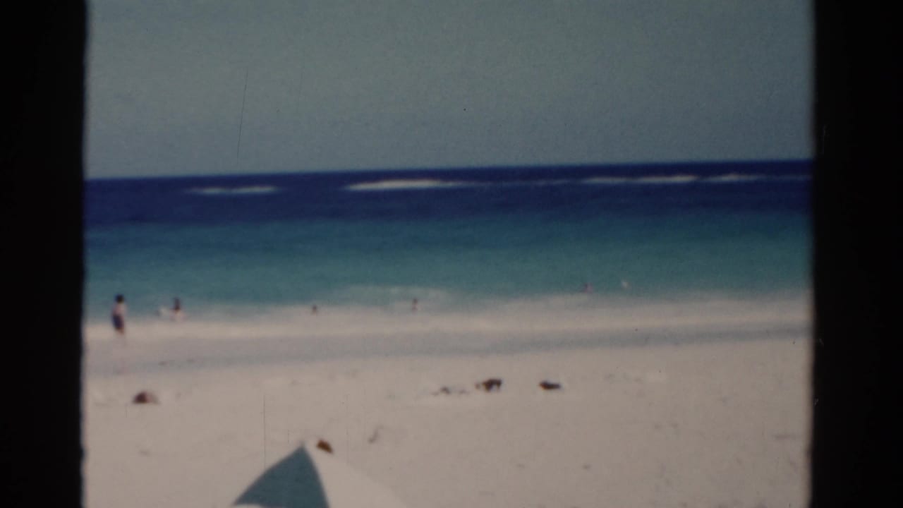 Wide shoreline with blue water, white sand, and small figures standing near the surf.
