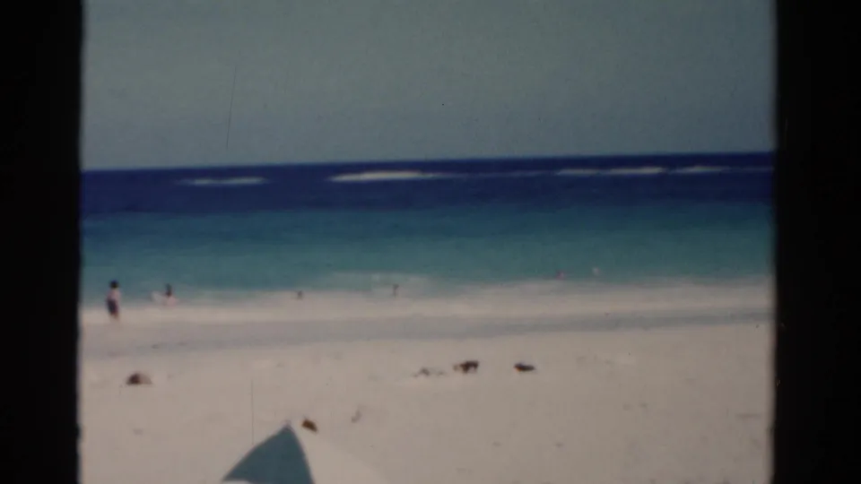 1975 Florida beach scene showing a bright shoreline, blue ocean bands, distant beachgoers, and soft 8mm film texture.