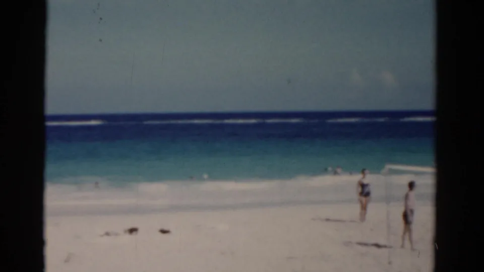 1975 Florida 8mm frame of people on a white beach near turquoise surf, with the sea horizon and film grain visible.
