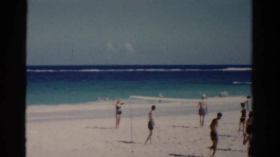 1975 Florida beach footage frame with beachgoers in swimwear, white sand, blue surf, and visible 8mm frame edges.