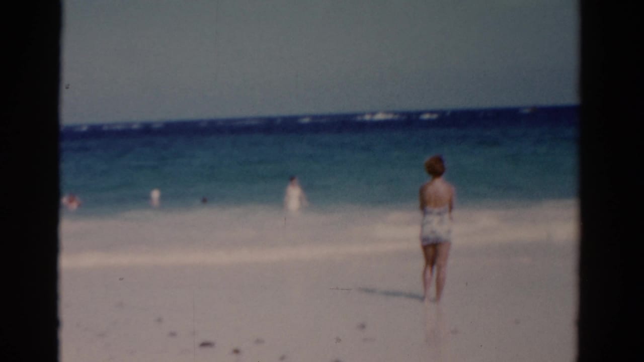 1975 Florida beach frame with a woman seen from behind on white sand, looking toward swimmers in the ocean.