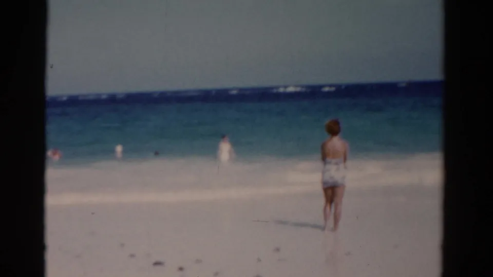 1975 Florida beach frame with a woman seen from behind on white sand, looking toward swimmers in the ocean.