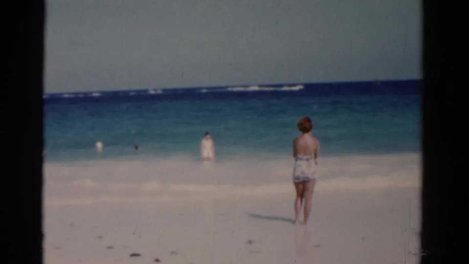 Soft 8mm 1975 Florida beach view of a woman from behind on sand with swimmers scattered across the surf line.