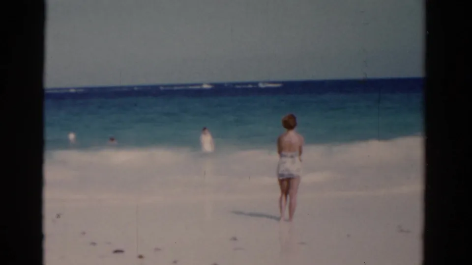 1975 Florida 8mm frame showing a woman standing on pale sand before blue-green ocean and distant bathers.