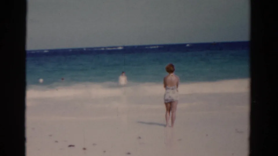 1975 Florida 8mm frame showing a woman in a patterned swimsuit standing on the beach and looking toward swimmers in the ocean.