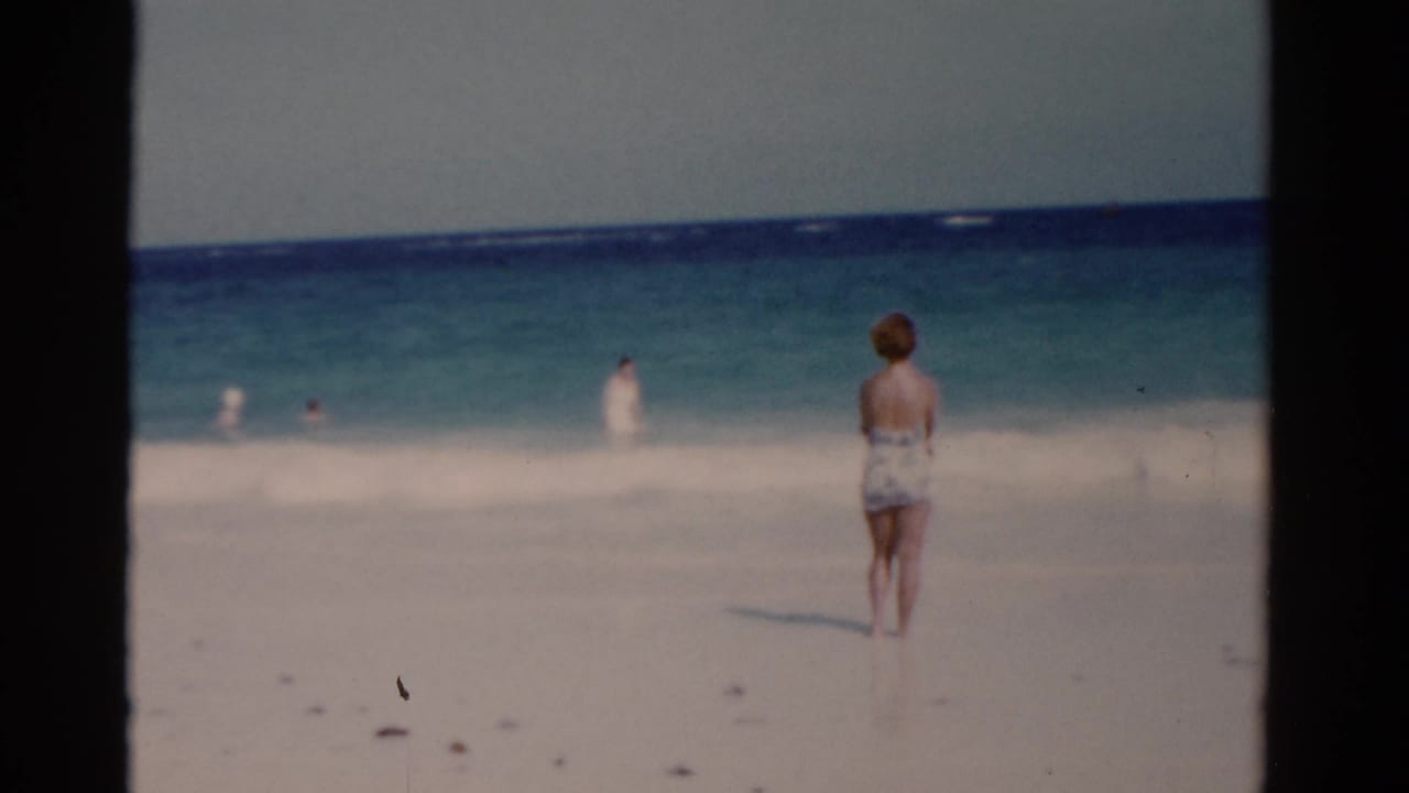 Woman on the beach faces the ocean while small figures swim in the waves.