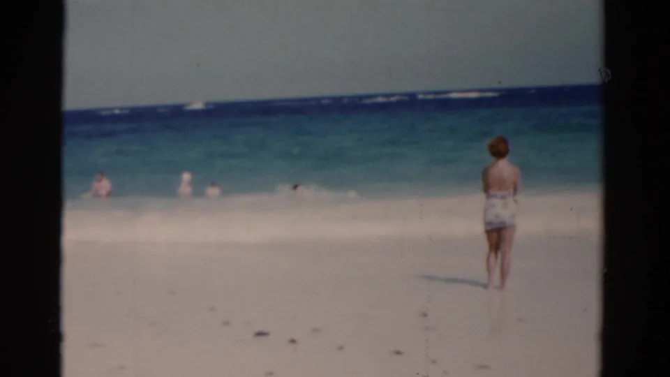 1975 Florida home-movie frame of a woman viewed from behind on a bright beach, with several swimmers visible in the shallow ocean.