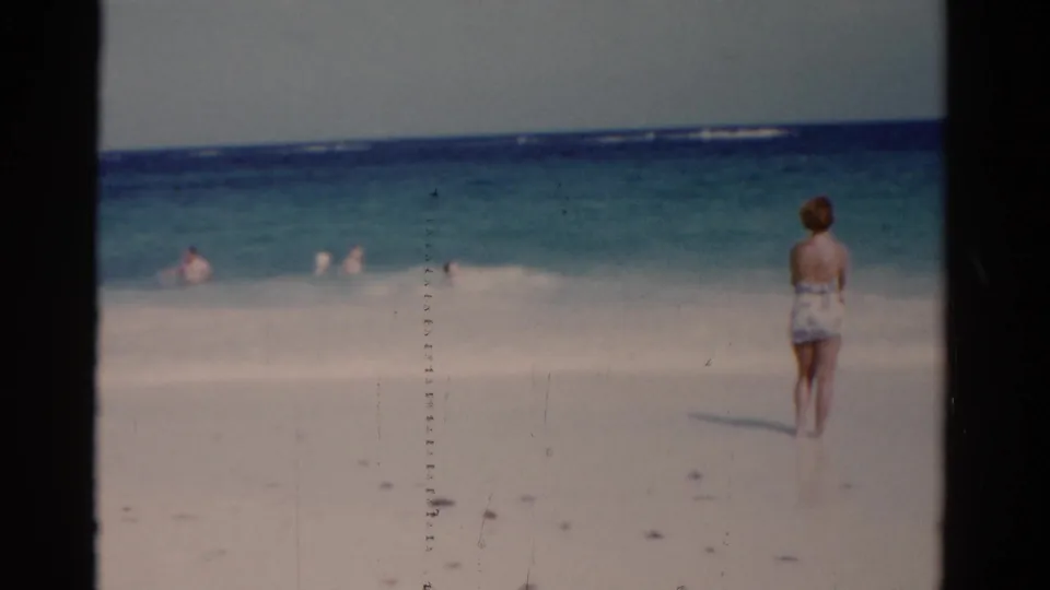 Faded 8mm frame from a 1975 Florida beach showing the woman at the right edge and swimmers scattered across the surf.