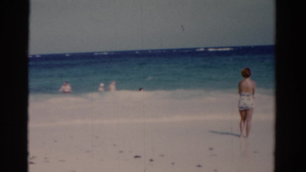 1975 Florida 8mm frame showing a woman in a patterned swimsuit facing the ocean as distant people play in the surf.