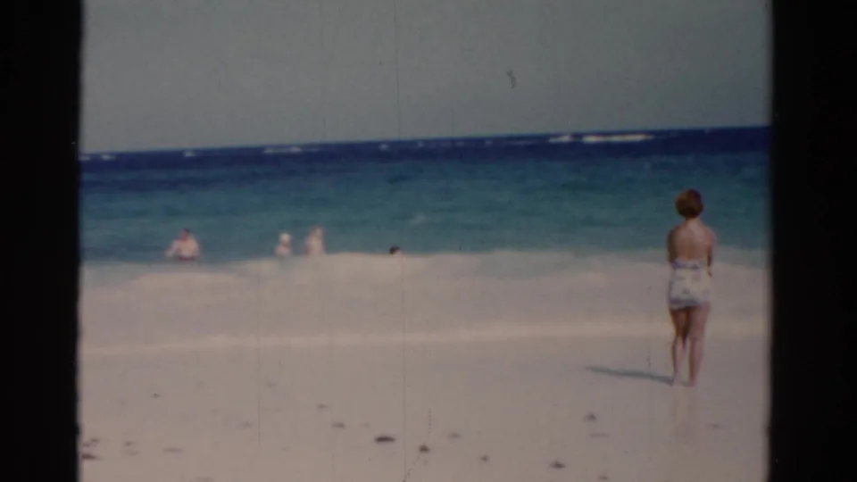 1975 Florida 8mm frame showing a woman in a patterned swimsuit facing the ocean as distant people play in the surf.