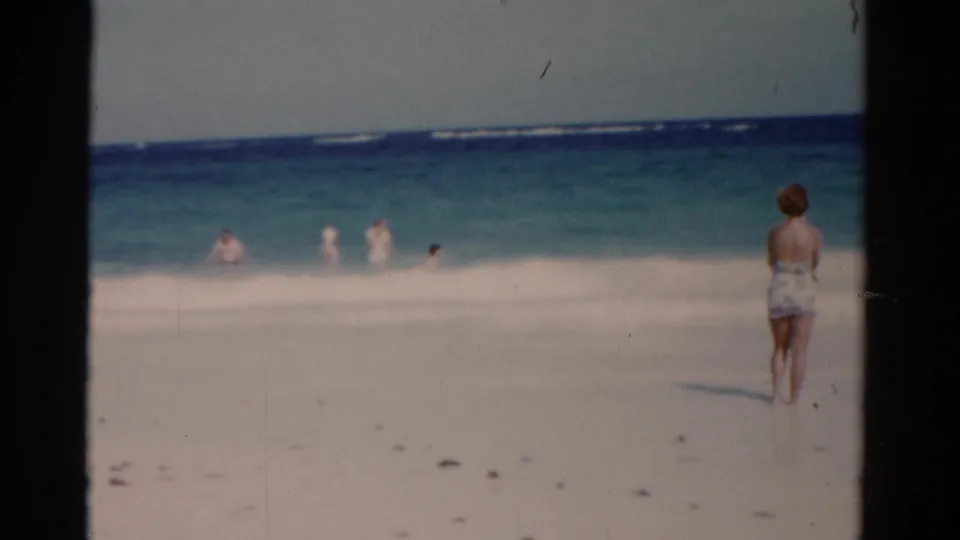 Soft 8mm frame from a 1975 Florida beach scene, with a woman on sand and several distant bathers in the waves.