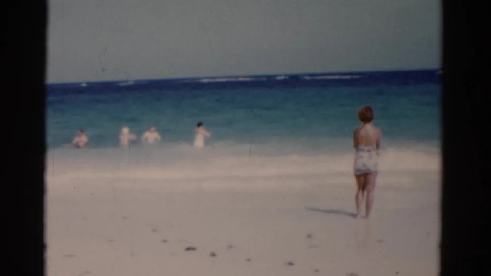 1975 Florida 8mm frame showing the woman at right, white beach sand, turquoise waves, and distant people in the water.