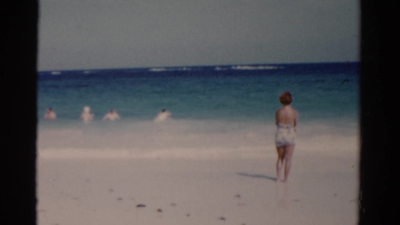 Woman in a swimsuit stands on a sandy beach facing the ocean.