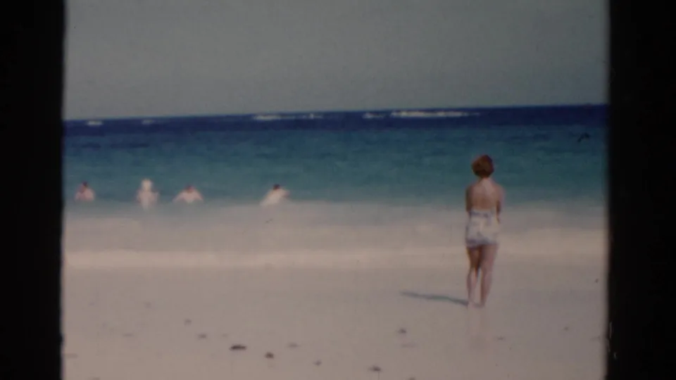 1975 Florida 8mm frame of a woman in a patterned swimsuit standing on white sand and looking toward the surf.