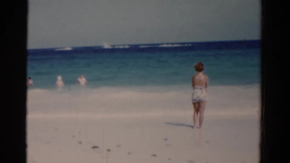 The woman remains near the Florida shoreline, facing the blue-green water and distant swimmers in this 8mm beach frame.