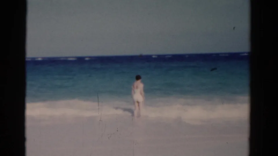 A figure in light swimwear stands in shallow Florida surf with blue water and white sand behind them.