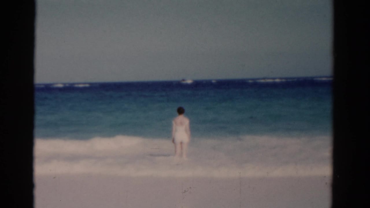Woman in a white swimsuit standing in shallow ocean waves near the horizon.