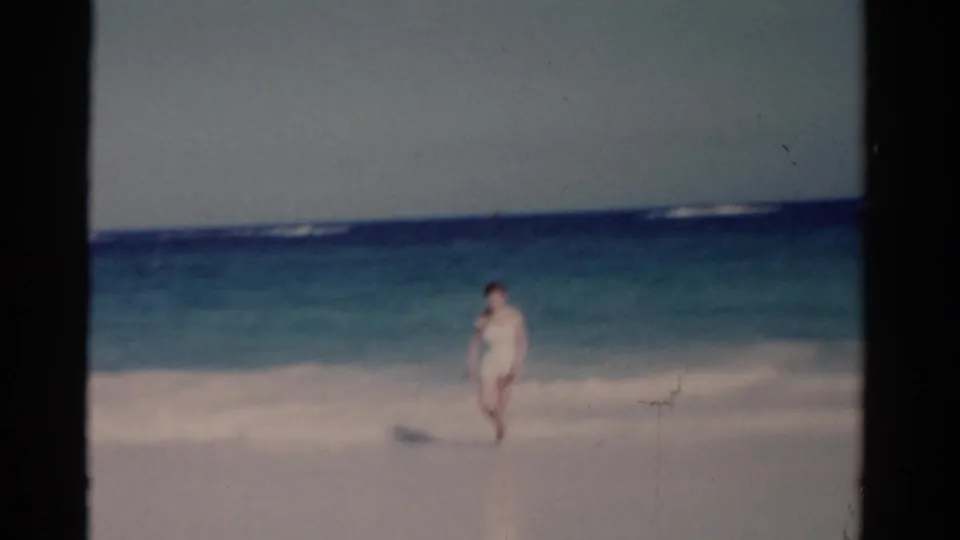 The woman walks out of the shallow Florida surf toward shore, with soft grain and fading color visible in the 8mm frame.