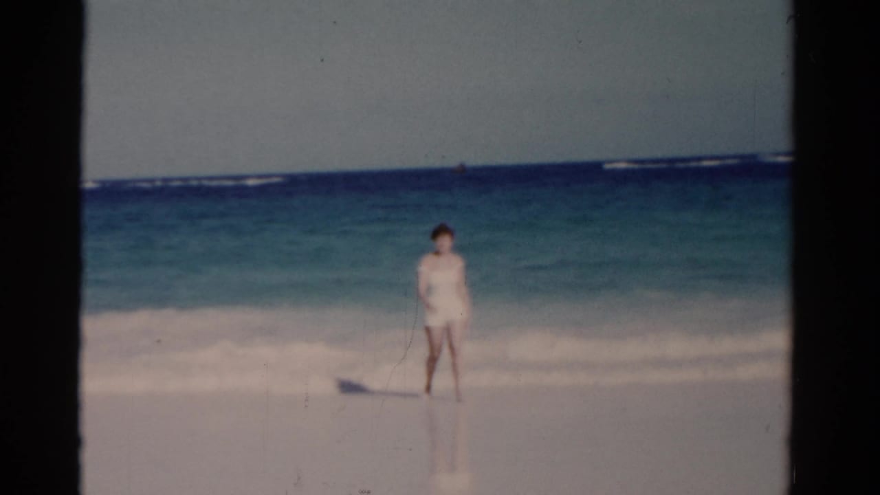 Woman in a white swimsuit standing near gentle surf on a sandy beach.