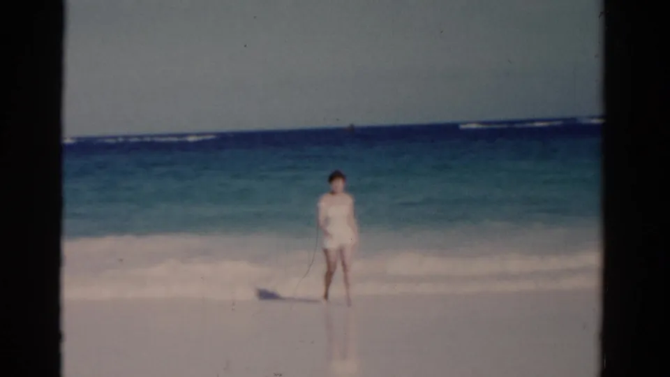 A woman in a white swimsuit stands at the shoreline in 1975 Florida home-movie beach footage, with blue water behind her.