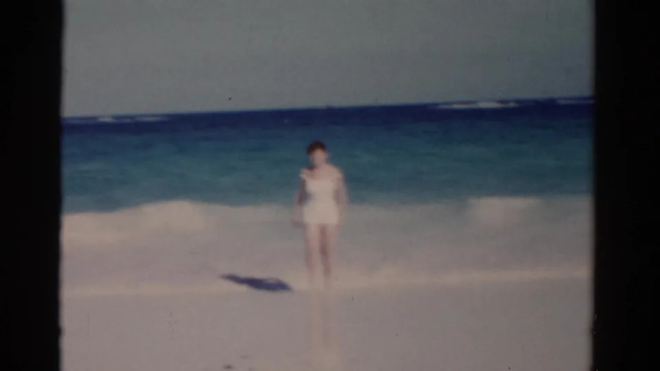 A woman walks on pale sand beside turquoise surf in Florida 1975 8mm footage, seen through soft home-movie focus.