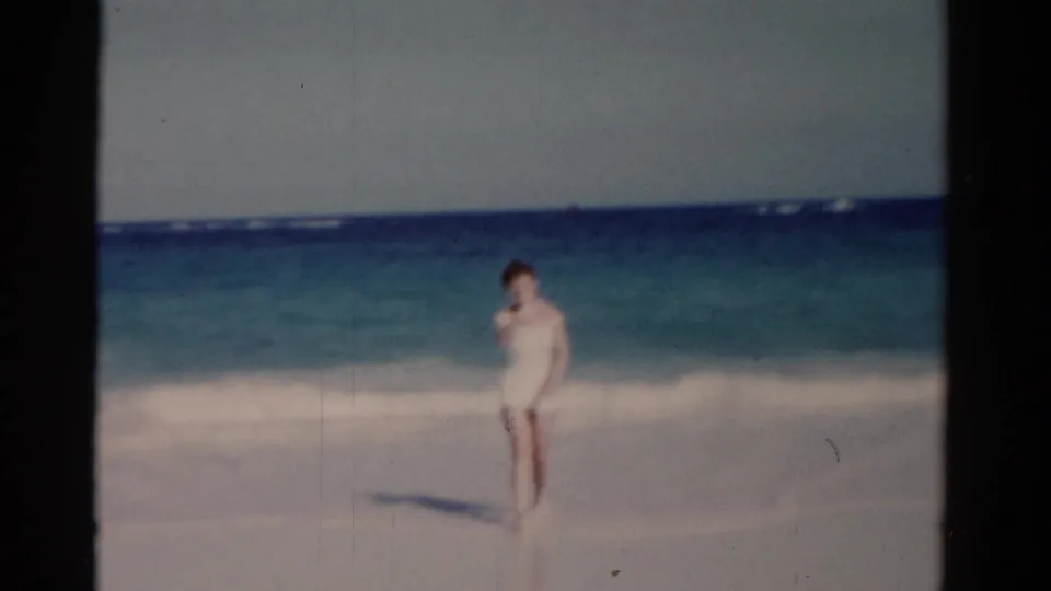 A woman approaches the camera from the surf line in 1975 Florida beach footage, with wet sand and low waves visible.