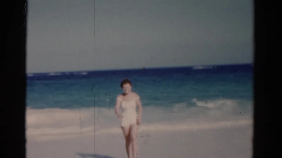 A woman in a white swimsuit walks toward the camera on a Florida beach in 1975, framed by surf, sand, and a hazy horizon.