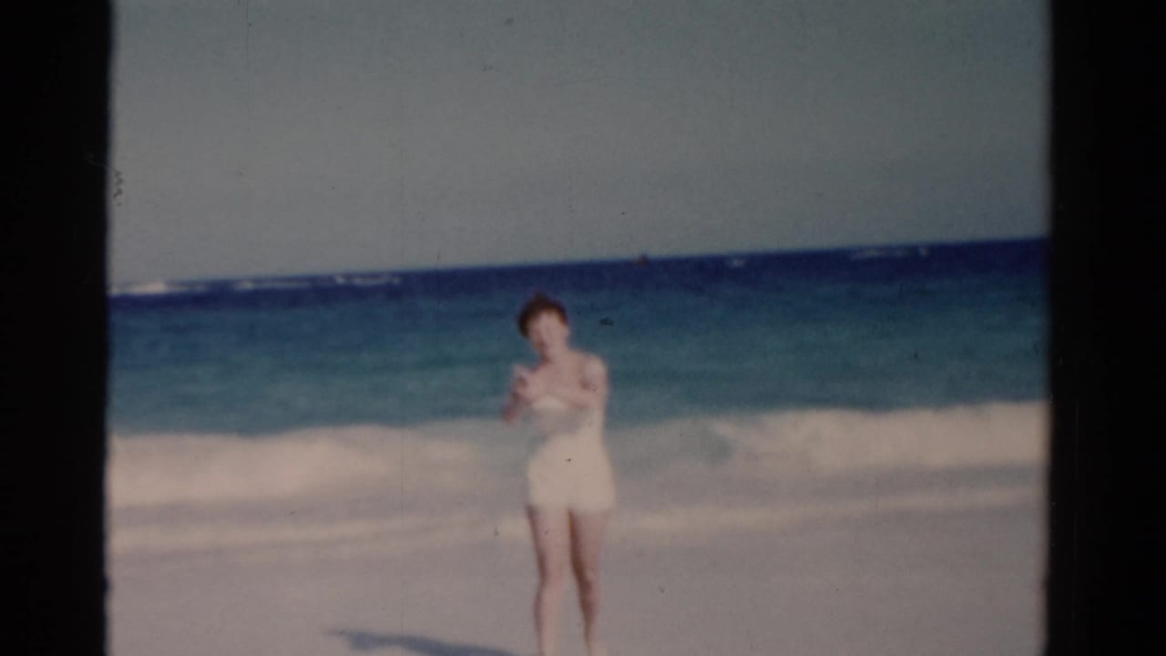 Woman in a white swimsuit standing on a sandy beach with ocean surf behind her.