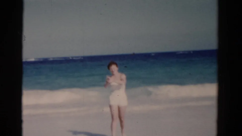 1975 Florida 8mm frame of a woman in a white swimsuit near the shoreline, with blue-green water and surf in the background.