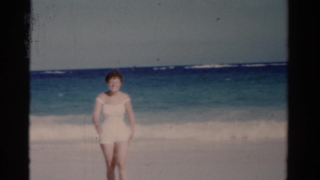 1975 Florida beach home-movie frame showing a woman facing the camera near rolling surf under a pale sky.