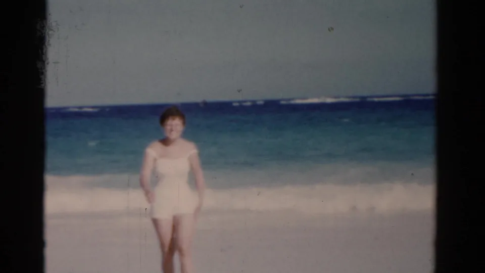 1975 Florida beach home-movie frame showing a woman facing the camera near rolling surf under a pale sky.