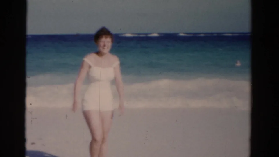 1975 Florida 8mm frame with a closer view of a woman walking toward the camera on sand, with ocean surf behind her.