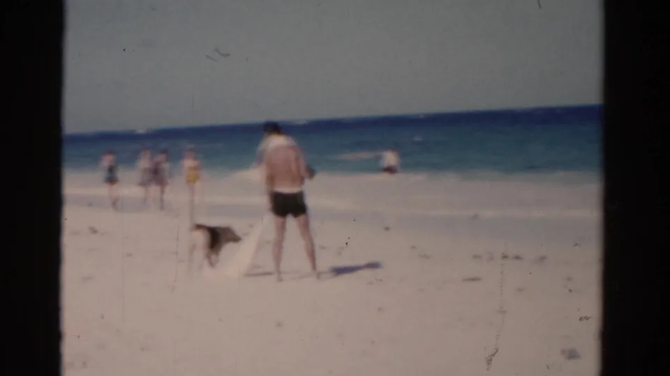 1975 Florida beach frame showing a man with a dog in the sand and several distant beachgoers near the shoreline.