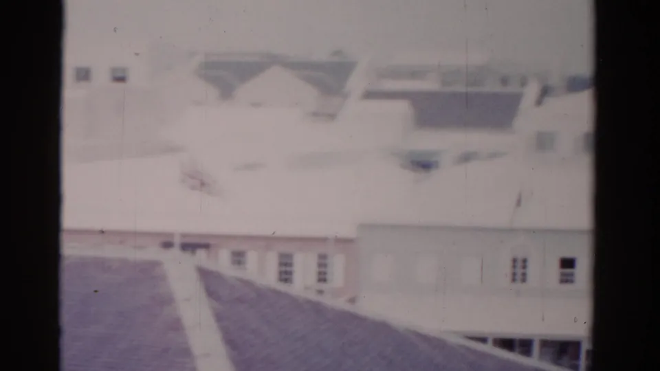 1975 Florida 8mm frame ending on pale rooftops and low-rise building walls with small windows and trees beyond.