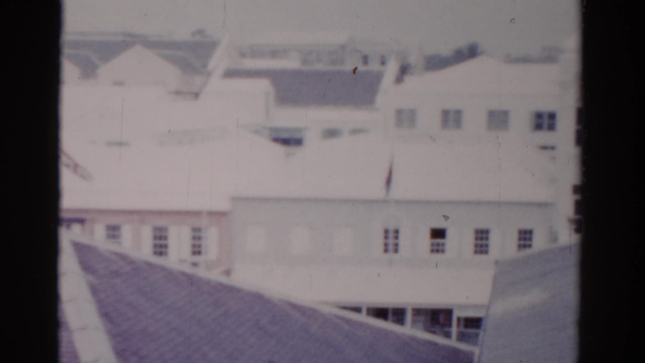 Soft 8mm view of pale buildings and rooftops seen through dark vertical window framing.