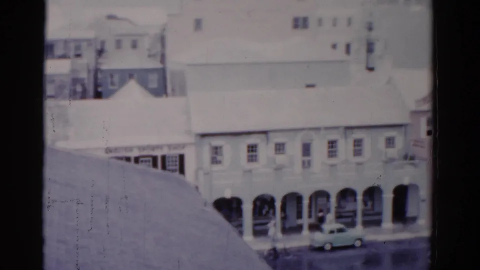 1975 Florida street view with arched ground-floor walkways, pale buildings, a small car, and soft 8mm film texture.