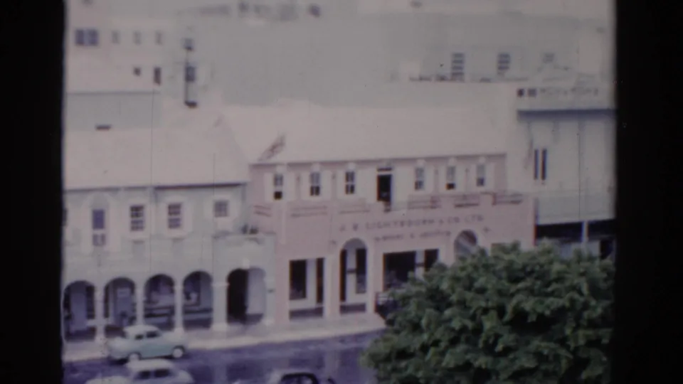 1975 Florida street frame with arched commercial facades, moving or parked cars, pedestrians, signage, and a leafy foreground tree.