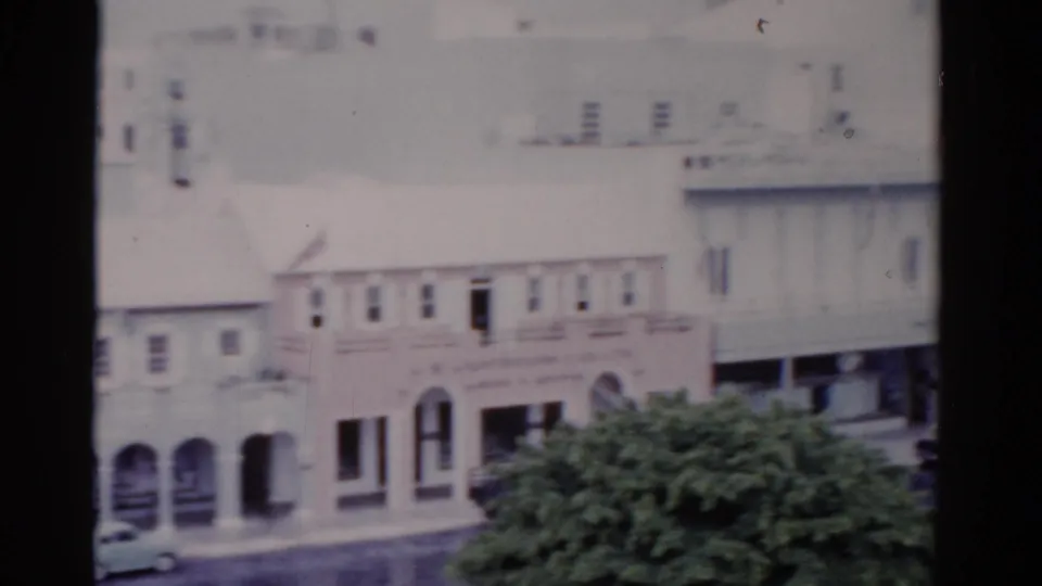 1975 Florida home-movie frame showing white low-rise commercial buildings, arched storefronts, leafy trees, and cars below.
