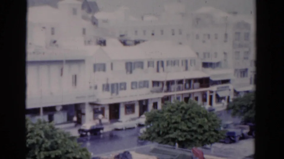 1975 Florida frame showing a low-rise commercial street with storefronts, trees, parked cars, and visible road traffic.
