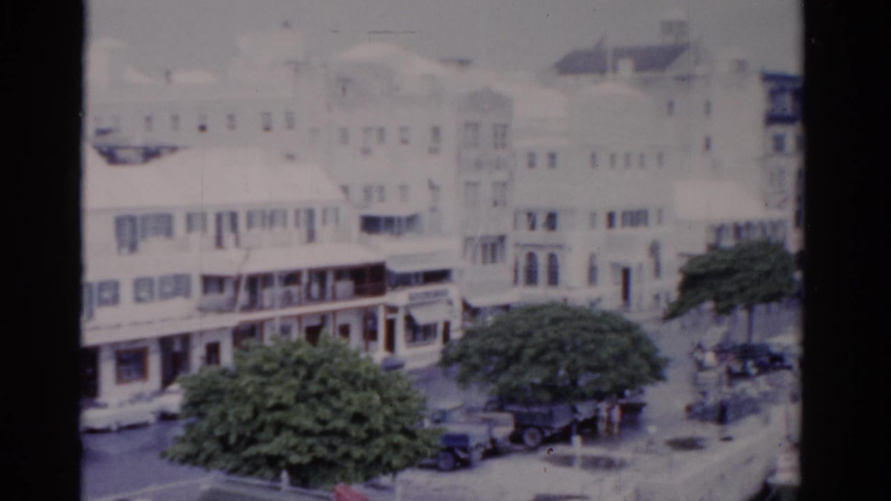 1975 Florida 8mm frame showing white waterfront buildings with balconies, leafy trees, parked cars, and distant pedestrians near the water.